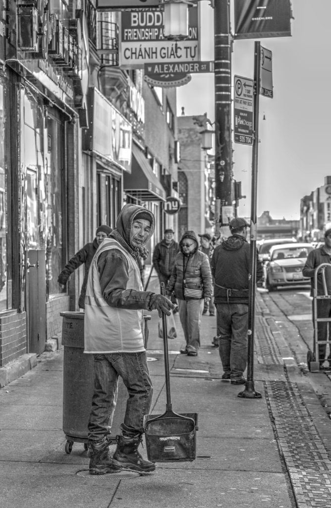 A street photography portrait of a Chinese man with a broom and garbage can, cleaning the sidewalks in Chicago Chinatown. Quest for Dirtman.