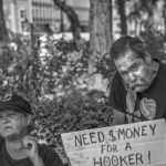 A recluse man holding up a cardboard sign that says, need money for a hooker.