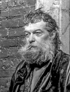 A black and white street portrait of a recluse man standing against a wall, smoking a cigarette.