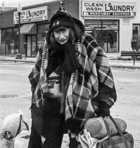 Street photo in black and white of an elderly woman walking across the street with bags of laundry.